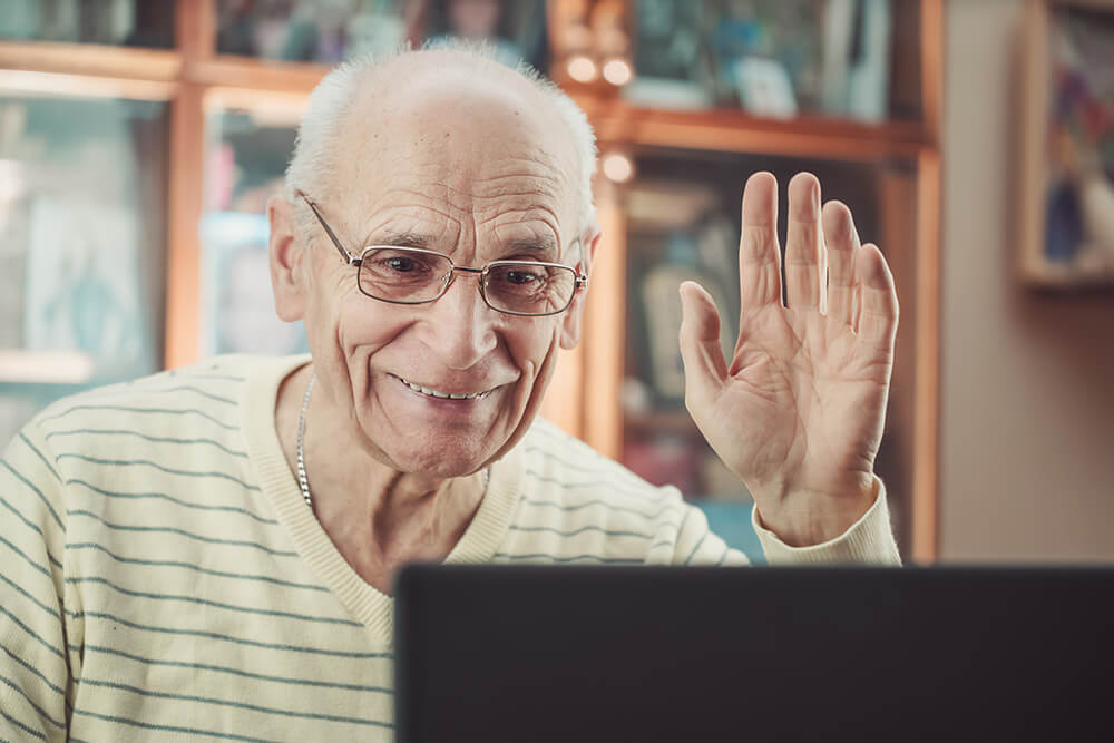 Senior man waving at the laptop, possibly with an Inclusee volunteer.