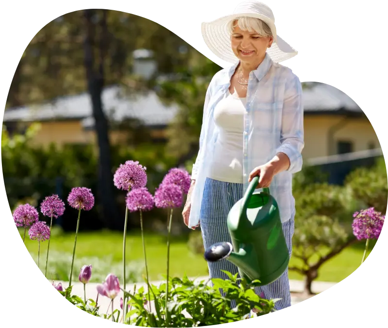 Elderly woman wearing a sun hat and gardening clothes, watering flowers in a garden.